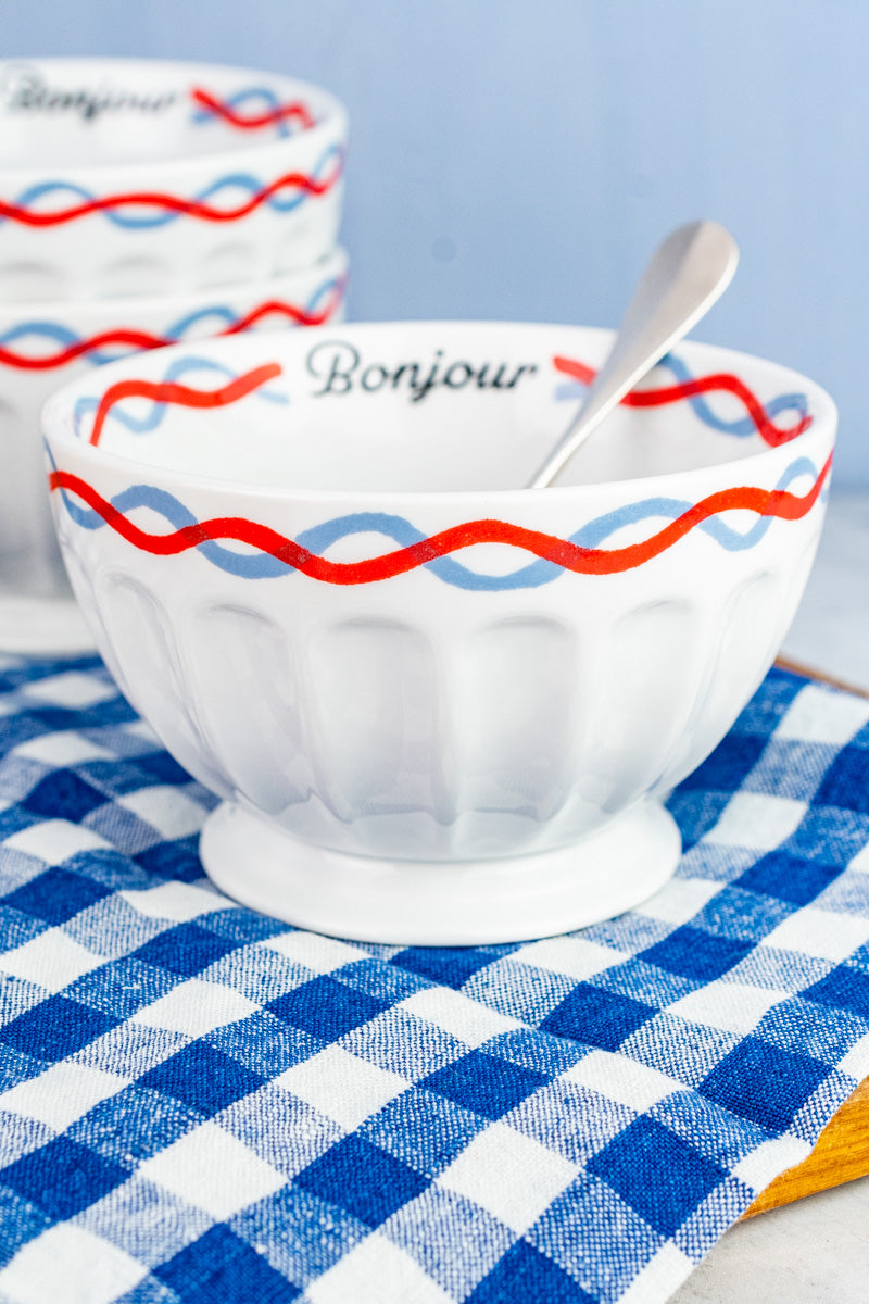 A white ceramic bowl with red and blue decorative stripes around the exterior rim, featuring the word 'Bonjour' on the interior rim, is placed on a blue and white checkered napkin. A silver spoon is also visible inside the bowl.